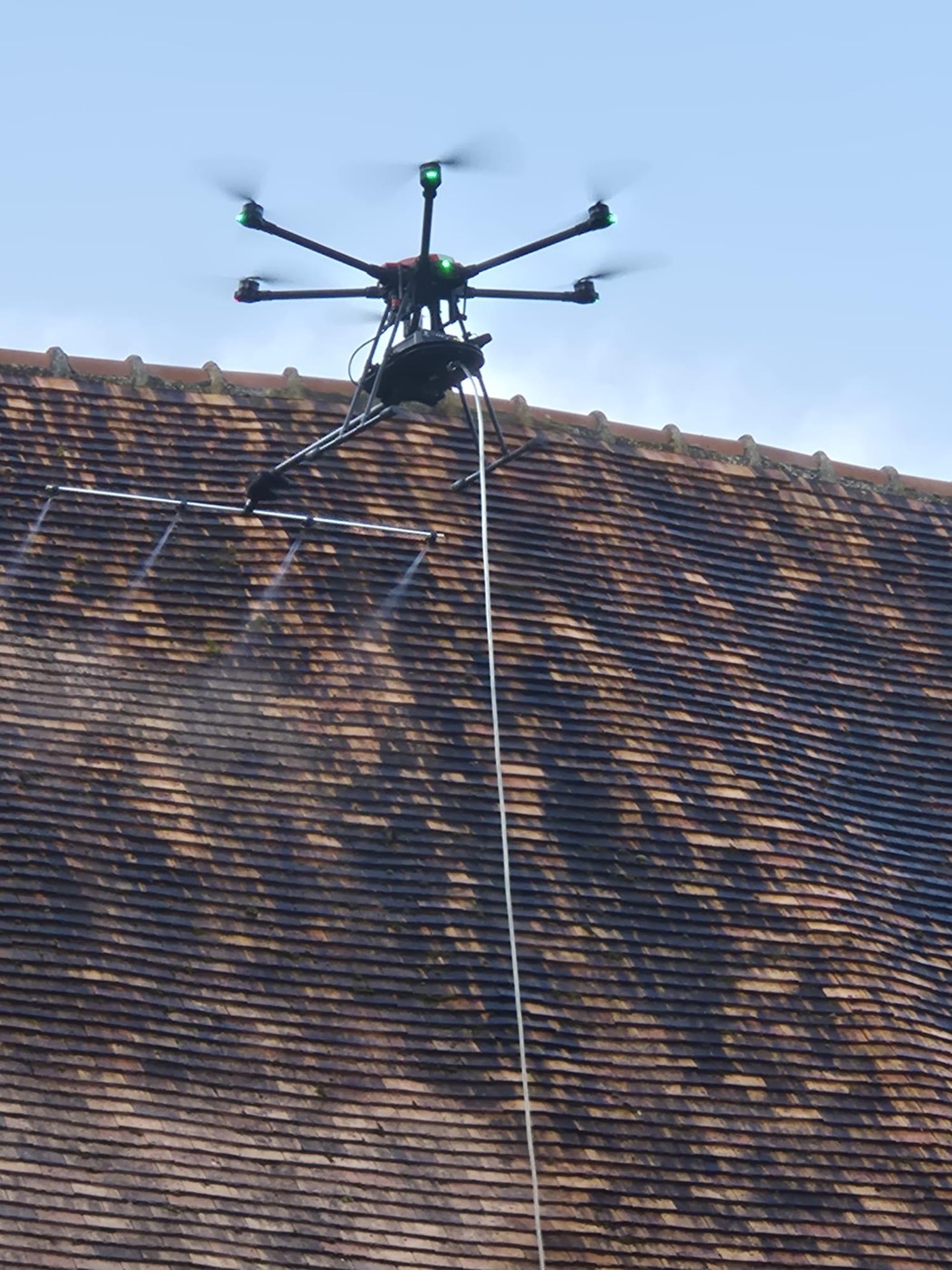 Un hexacoptère Altipur équipé d’une rampe de pulvérisation traite une toiture en tuile fortement encrassée ; la bande déjà traitée apparaît plus claire.