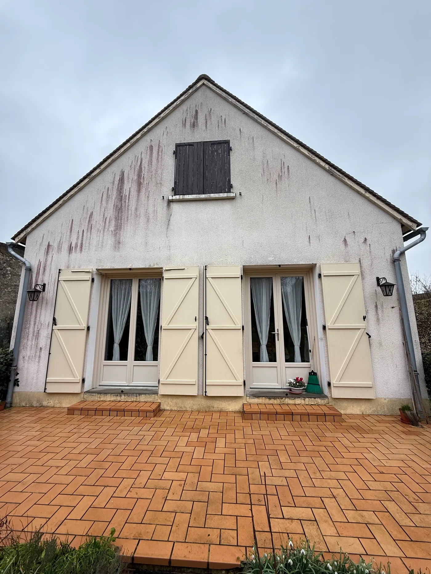 La façade d’une maison à volets, envahie de traînées rosées d’algues sur tout l’enduit clair.